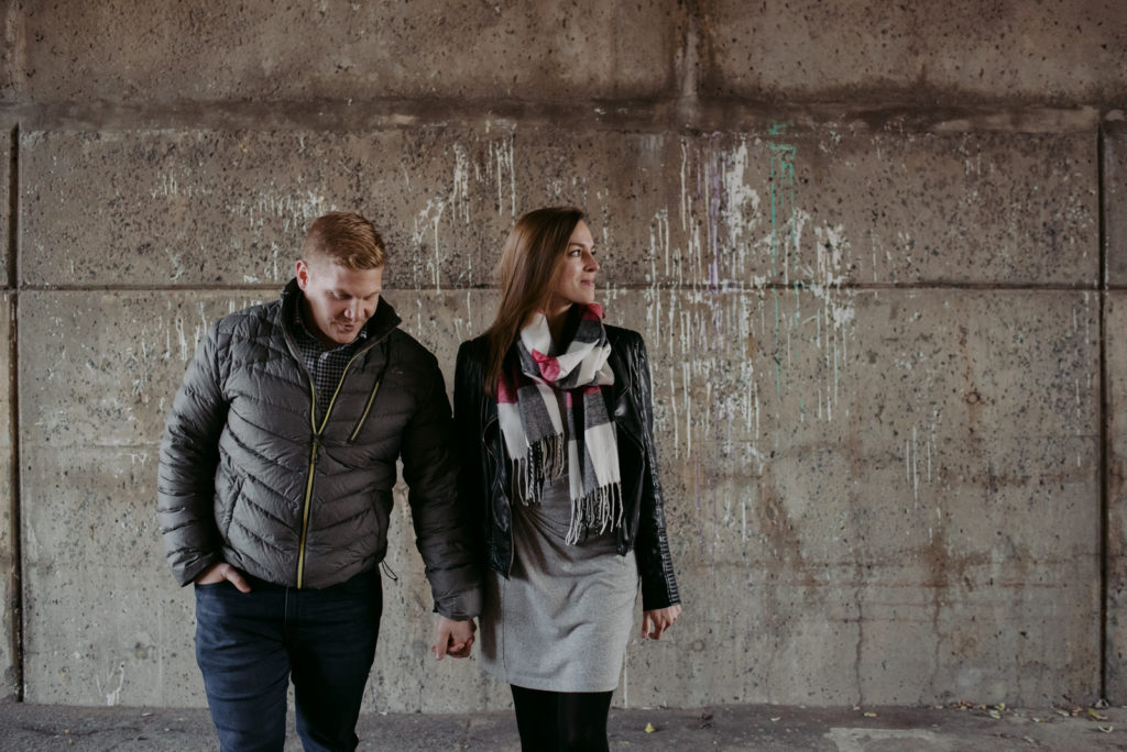 engaged couple holding hands and walking under concrete bridge
