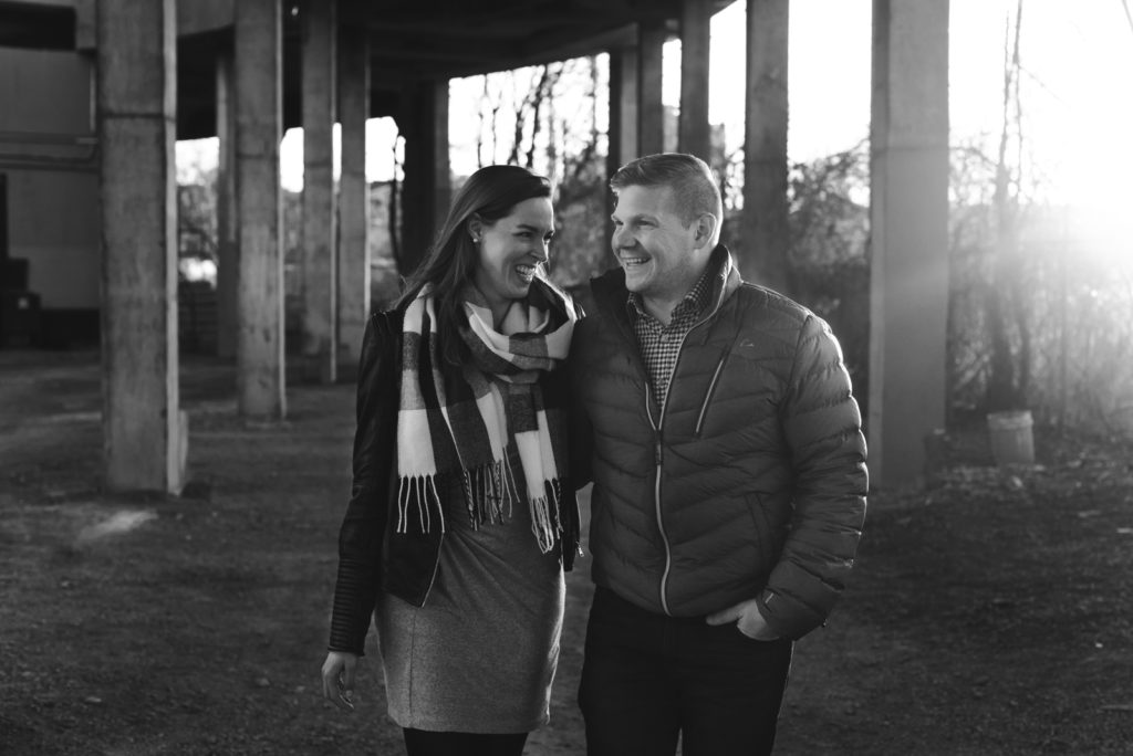 engaged couple laughing with arms around each other underneath a bridge at sunset