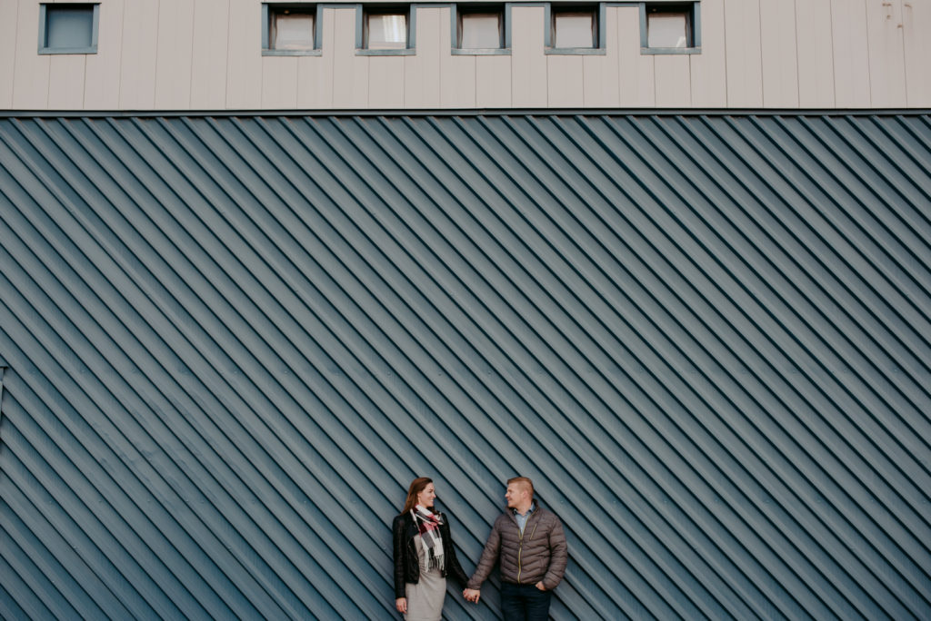 engaged couple against blue wall holding hands