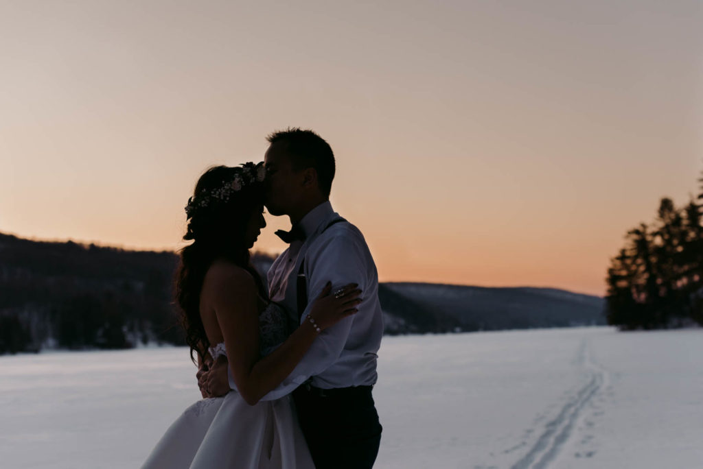 groom kissing the bride's forehead on a frozen lake at sunset