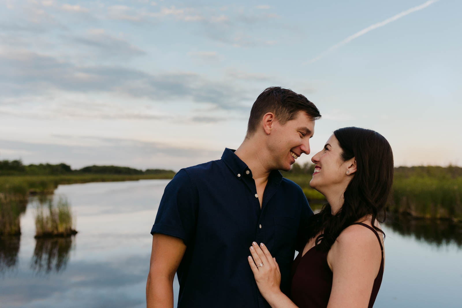 engaged couple smiling at each other by the water at sunset