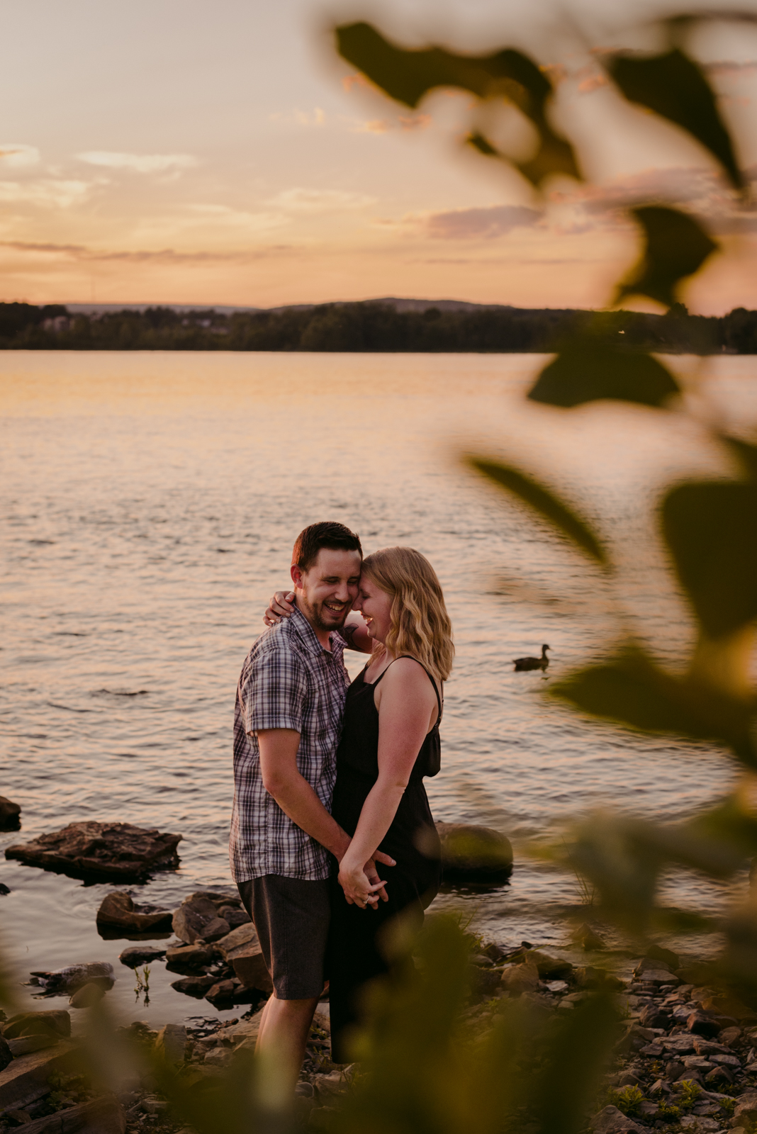 engaged couple cuddling and laughing by the water at sunset