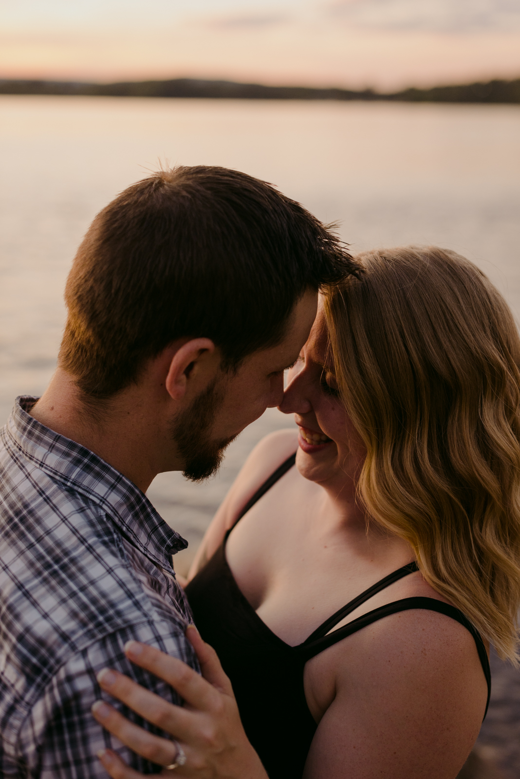 engaged couple with foreheads together laughing at sunset by the water