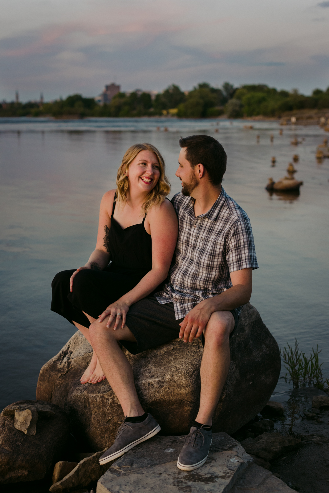 engaged couple sitting on the rocks by the water at sunset laughing