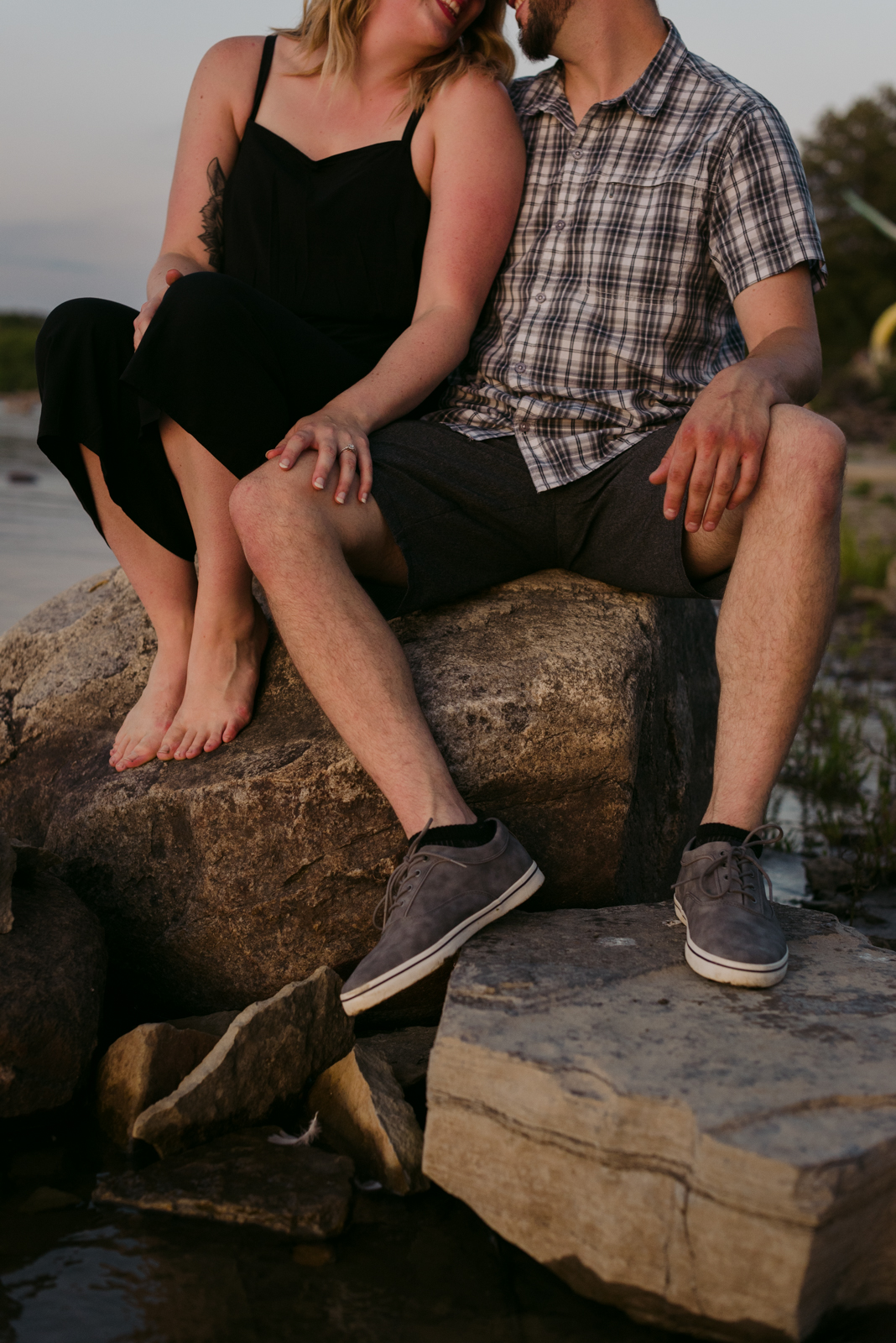 engaged couple sitting on the rocks by the water at sunset