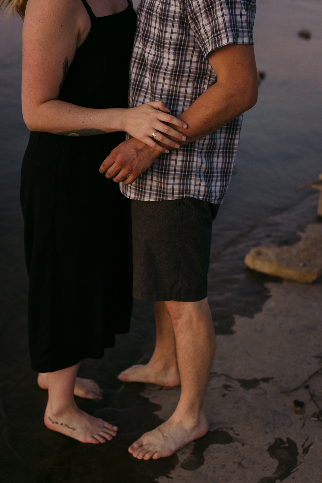 engaged couple with arms around one another and feet in the water