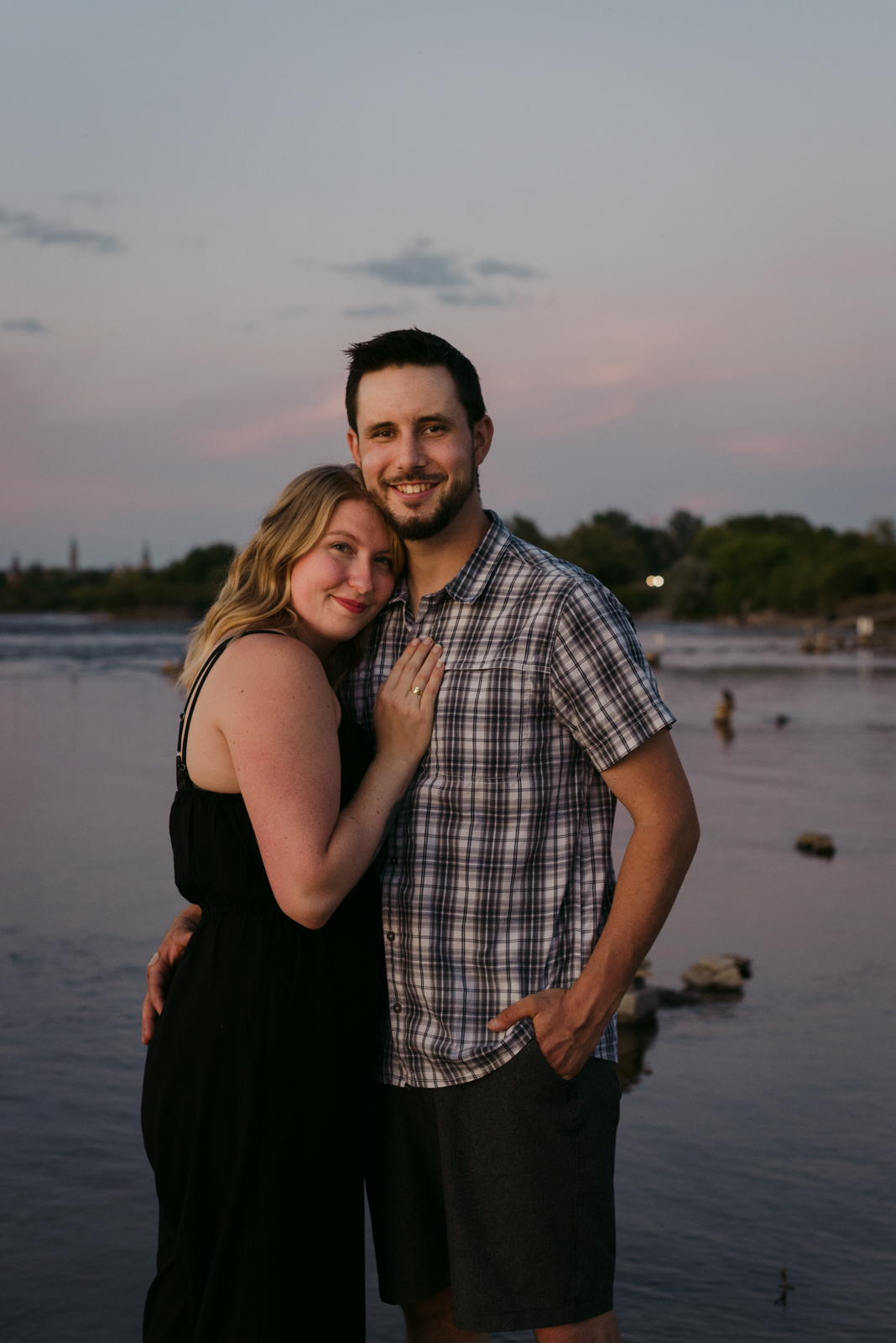 engaged couple smiling at the camera by the water at sunset
