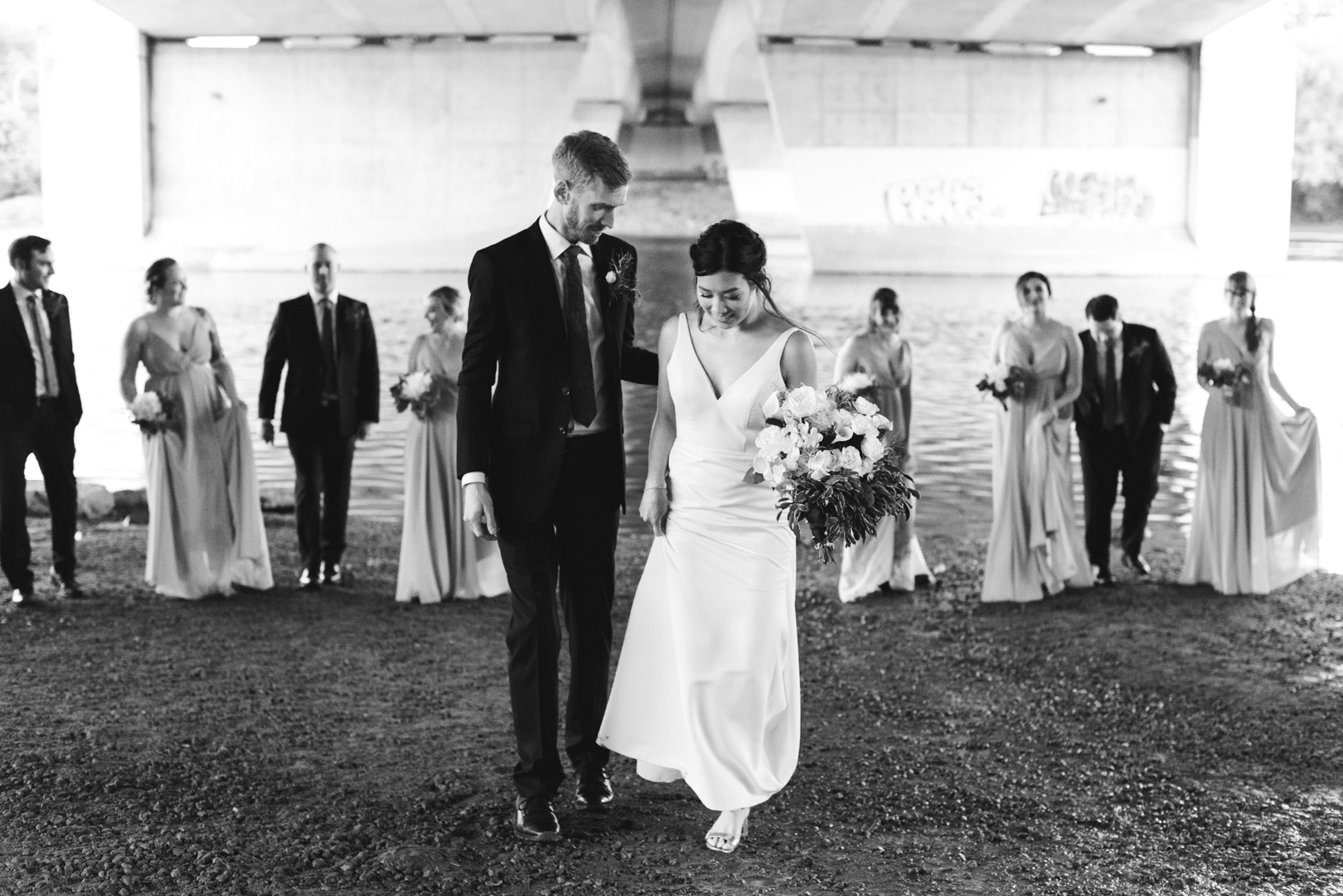 bride and groom walking with wedding party under bridge in black and white