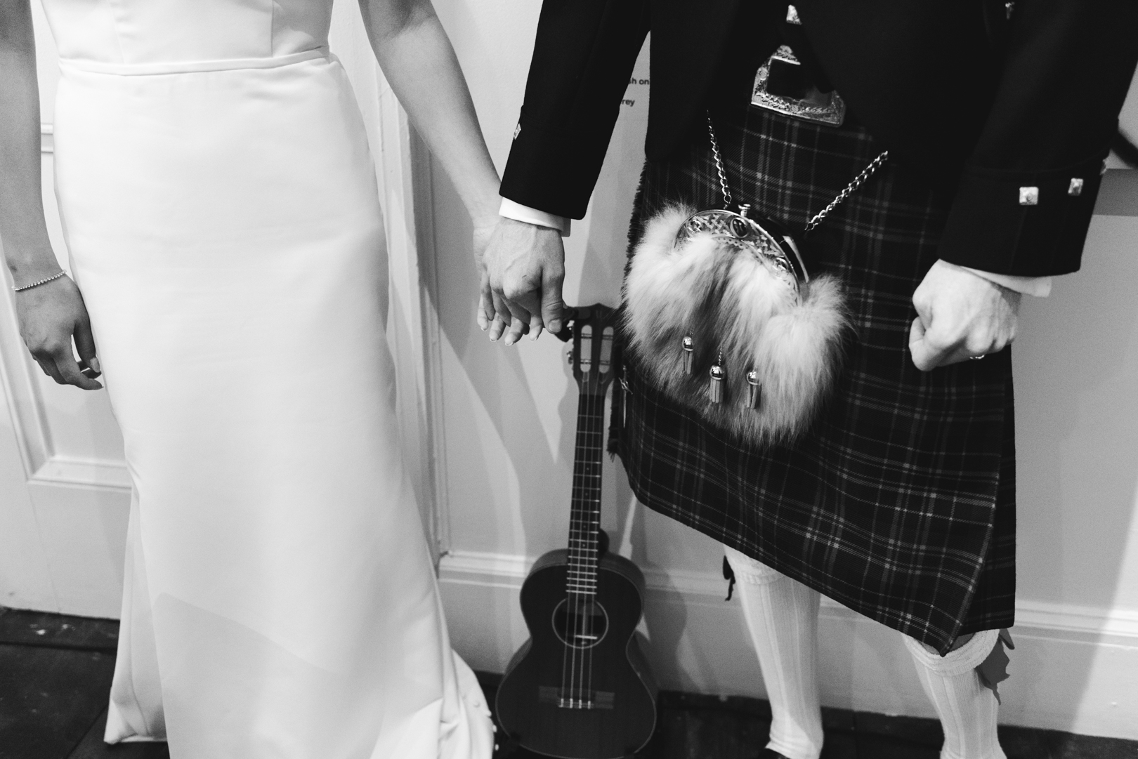 bride and groom wearing kilt holding hands with ukulele between them in black and white