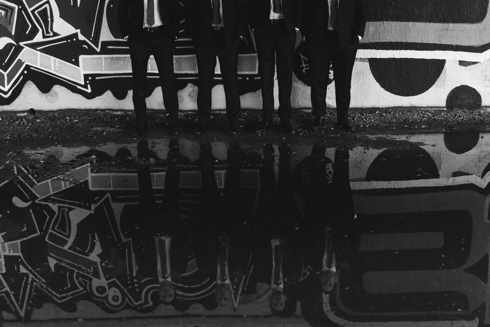 groom and groomsmen reflection in a puddle of water underneath a bridge with graffiti