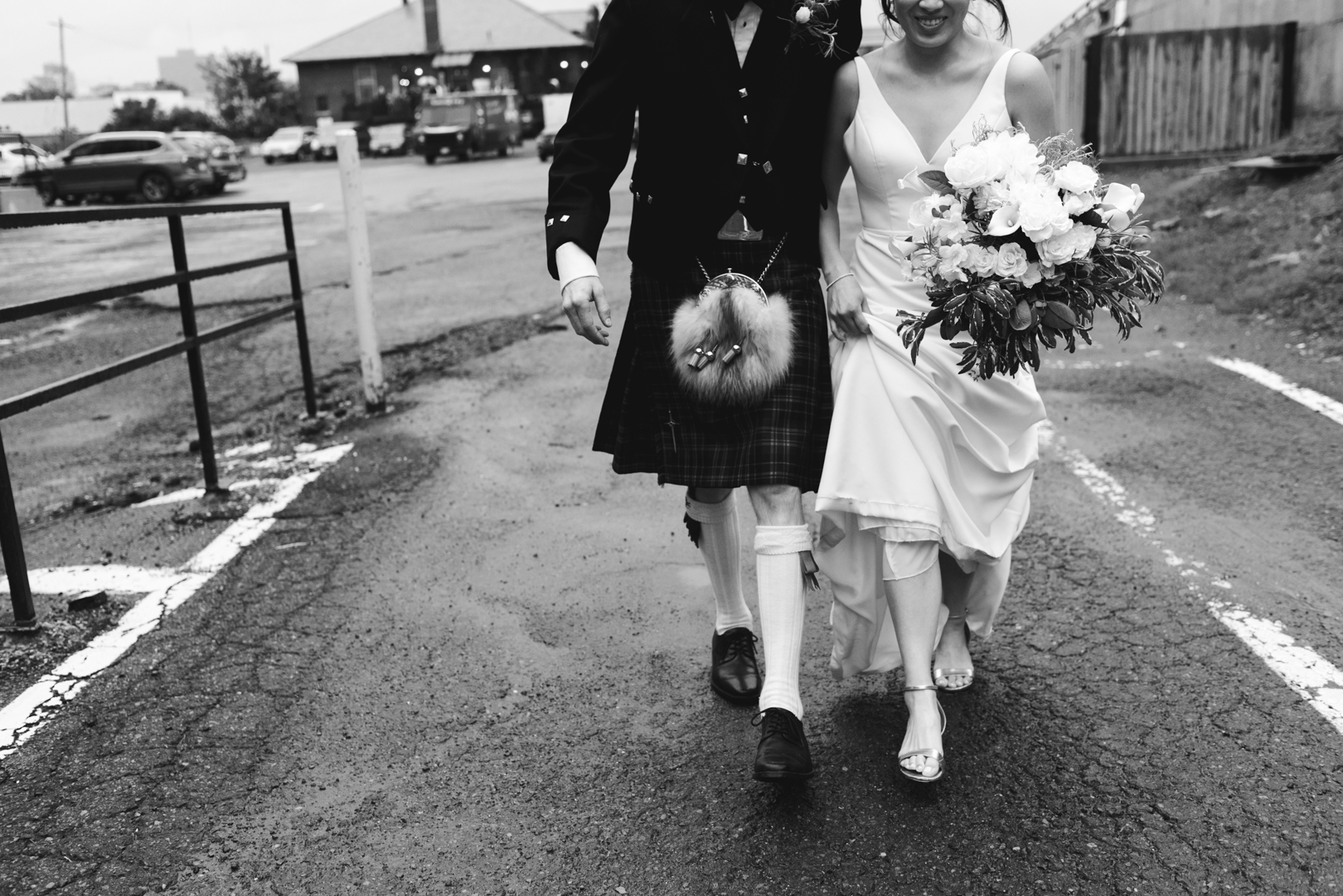 groom wearing kilt walking through parking lot with his bride in black and white