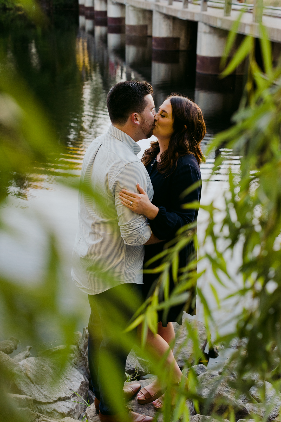 engaged couple kissing by the water underneath willow tree at sunset