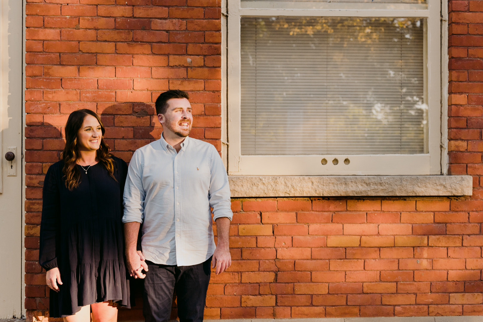 engaged couple holding hands against red brick building looking into the sunset