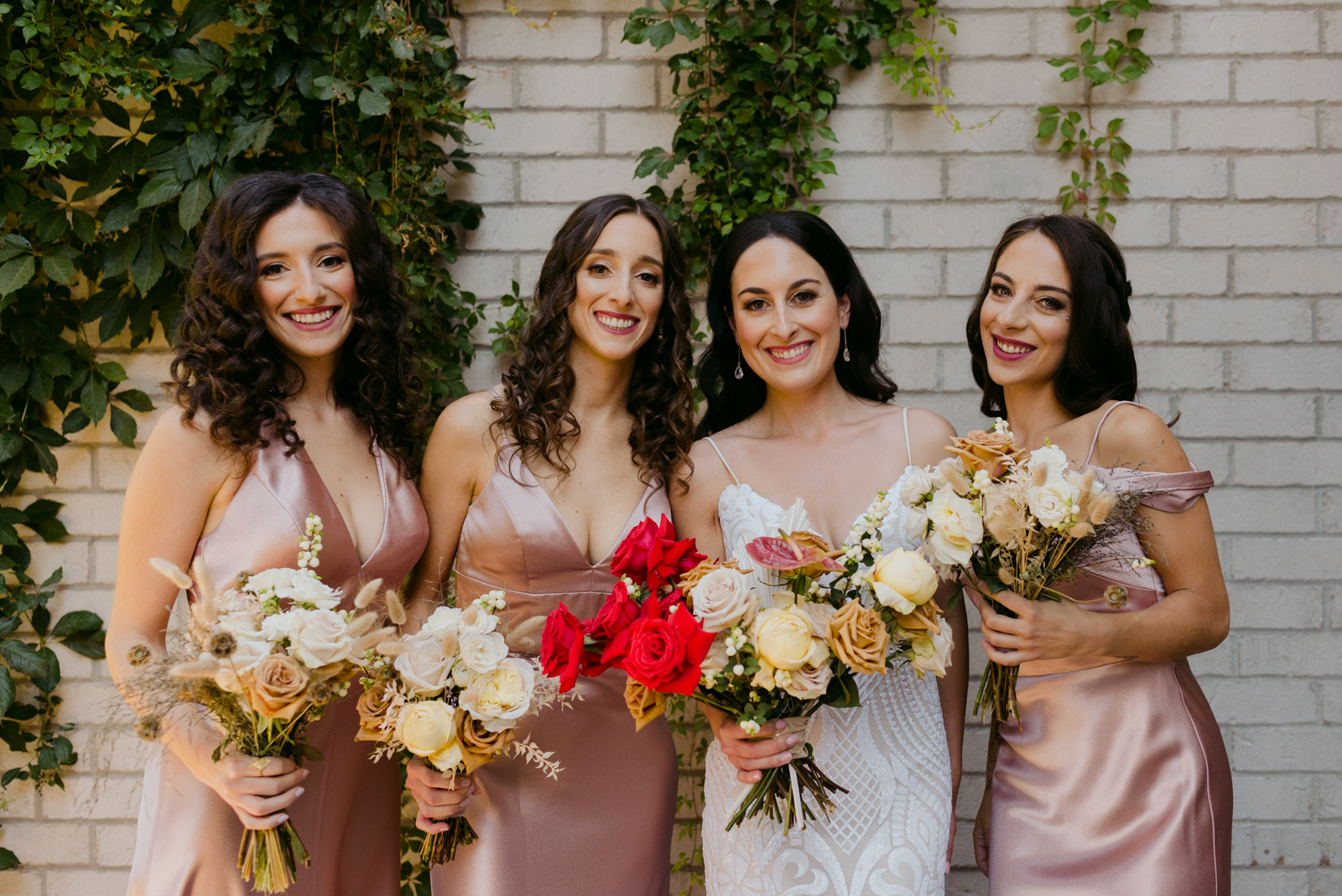 bride and bridesmaids in rose gold outside against white brick wall with vines