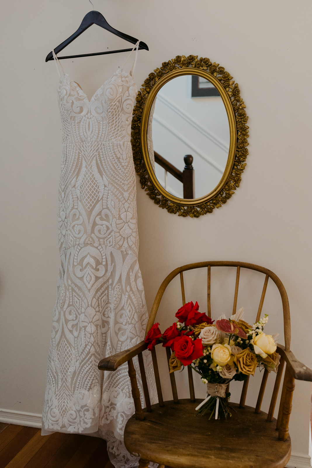 wedding dress hanging next to mirror with bouquet on vintage chair