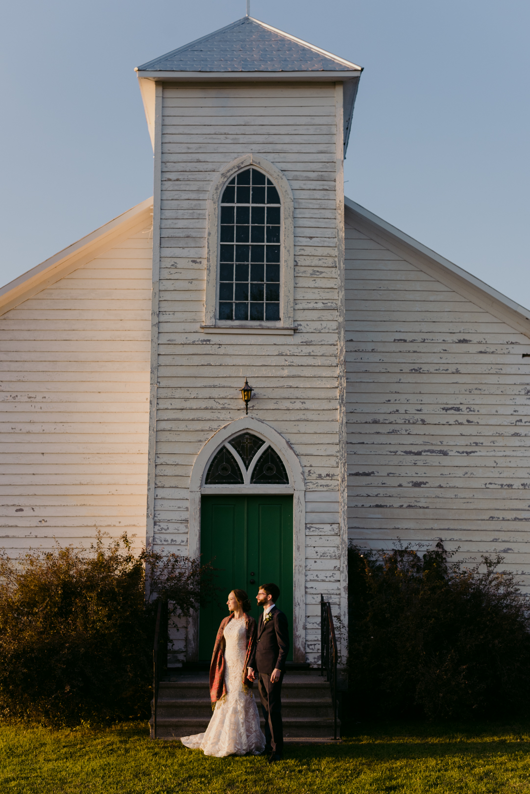 bride and groom standing in front of church looking into the sunset