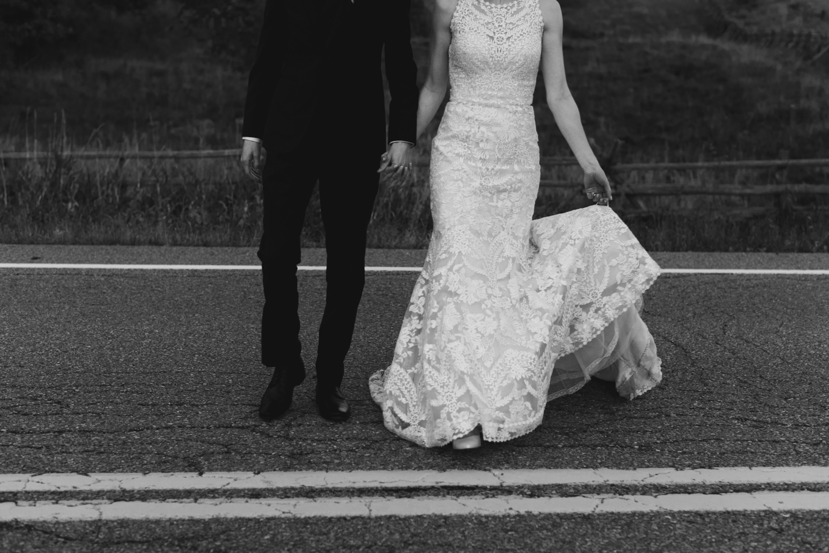 bride holding her train and her groom's hand as they walk across a country road in black and white
