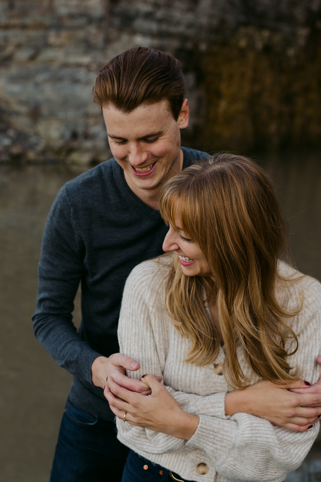 engaged couple with arms around each other laughing by the water