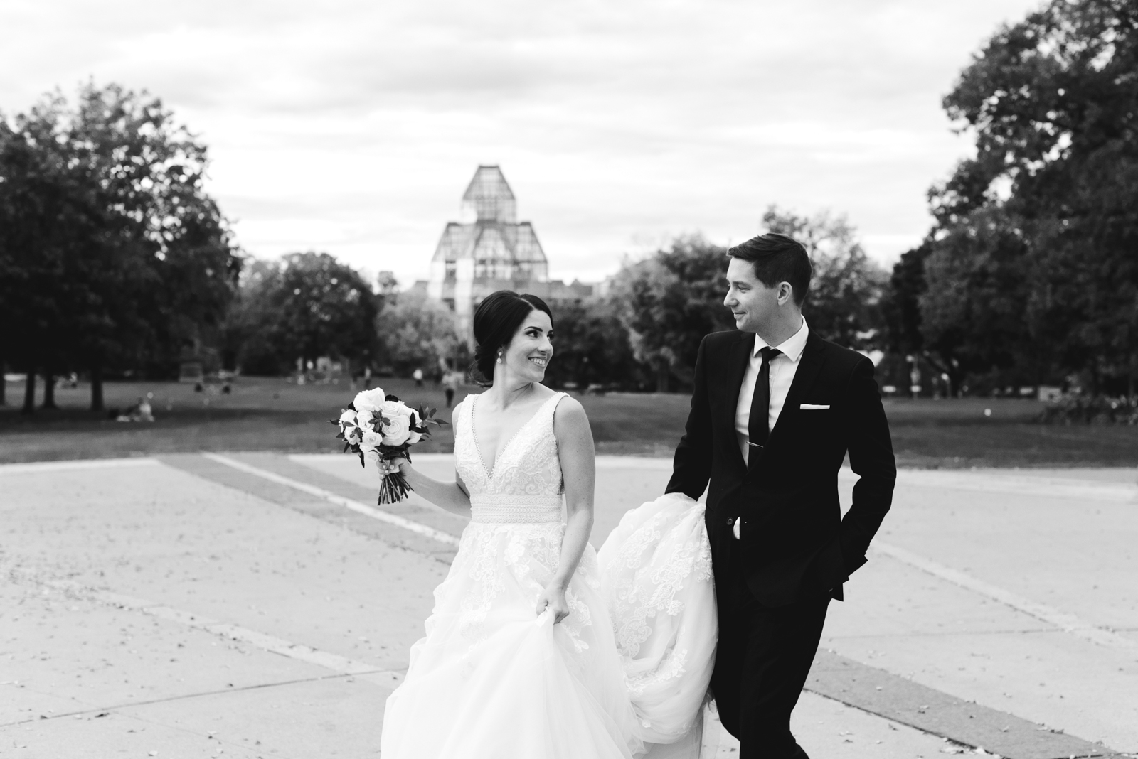 groom holding the bride's train as they walk through Majors Hill Park with National Gallery of Canada