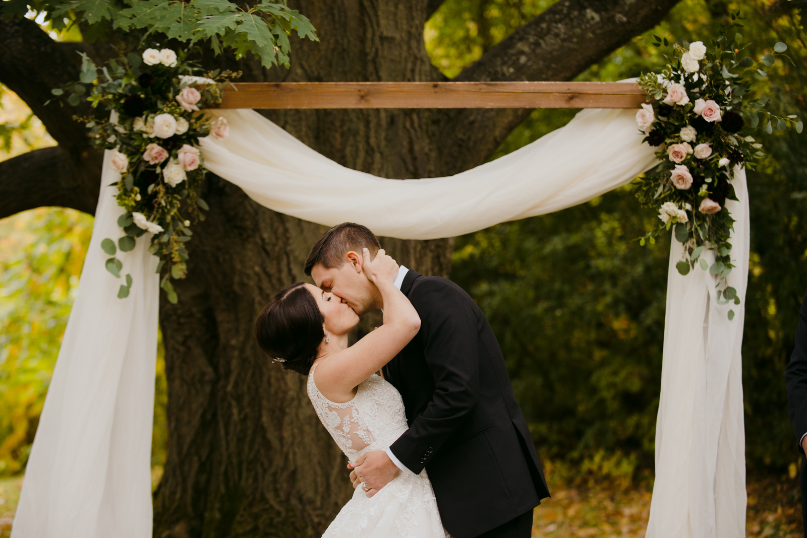 bride and groom first kiss under wooden arch during outdoor wedding ceremony
