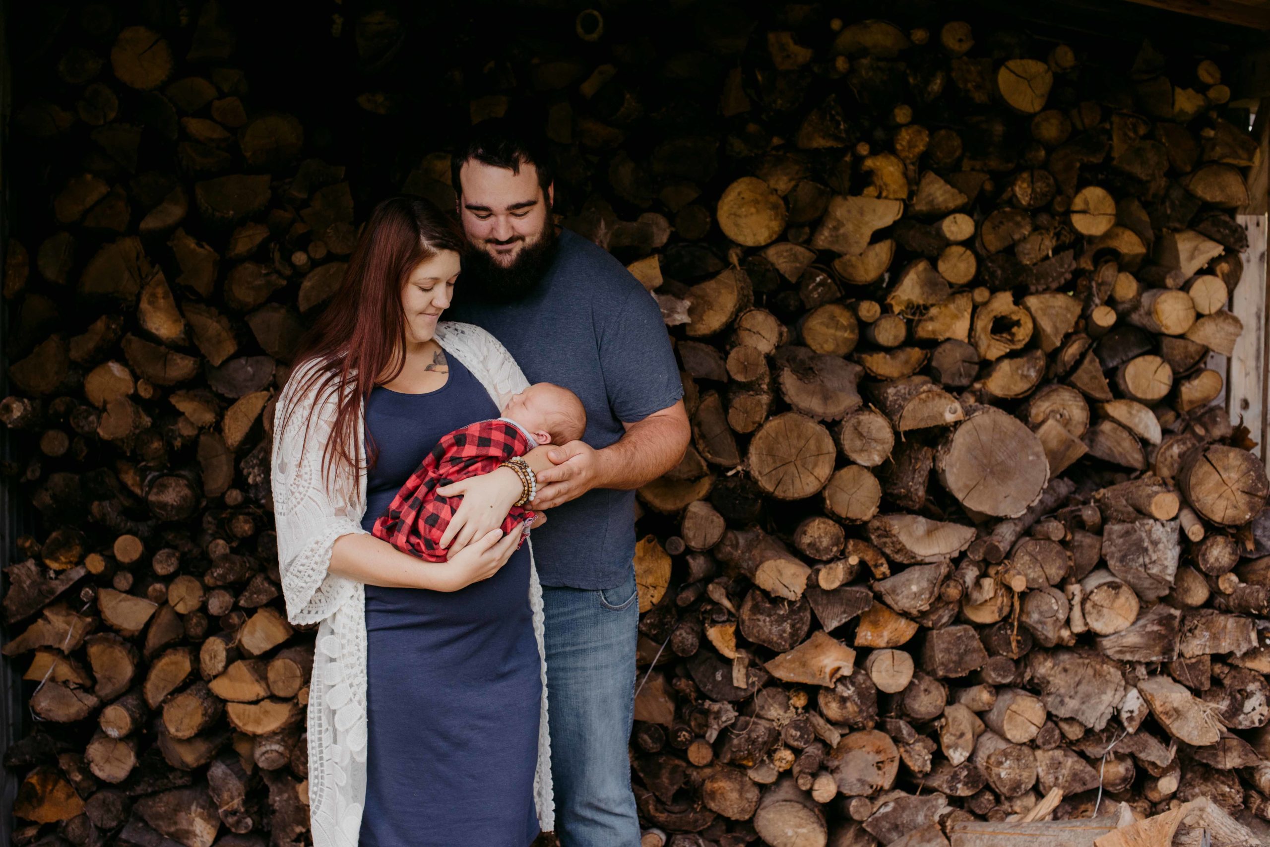mom and dad with baby boy in front of stack of wooden logs