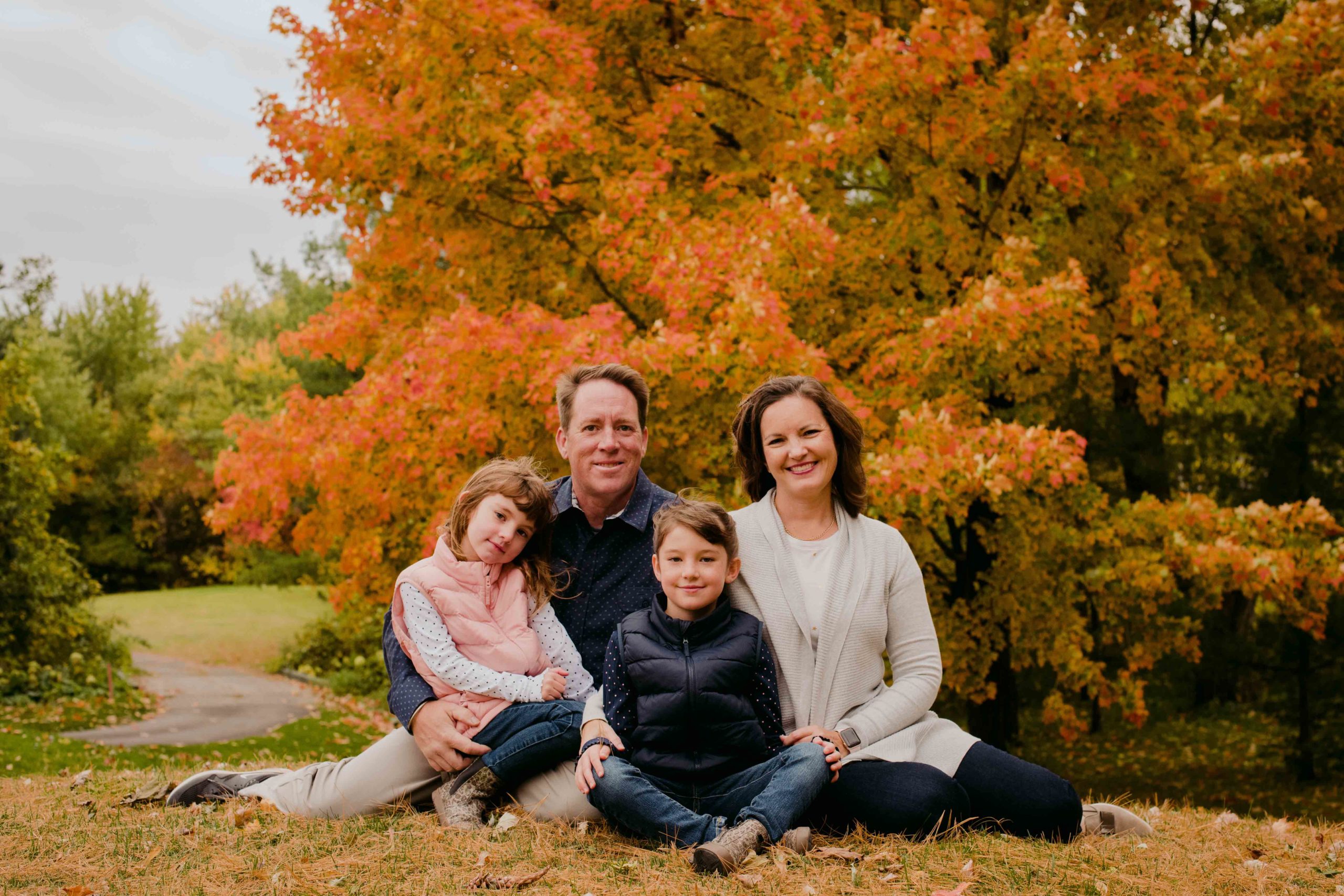 family of four sitting on the leaves in front of autumn tree