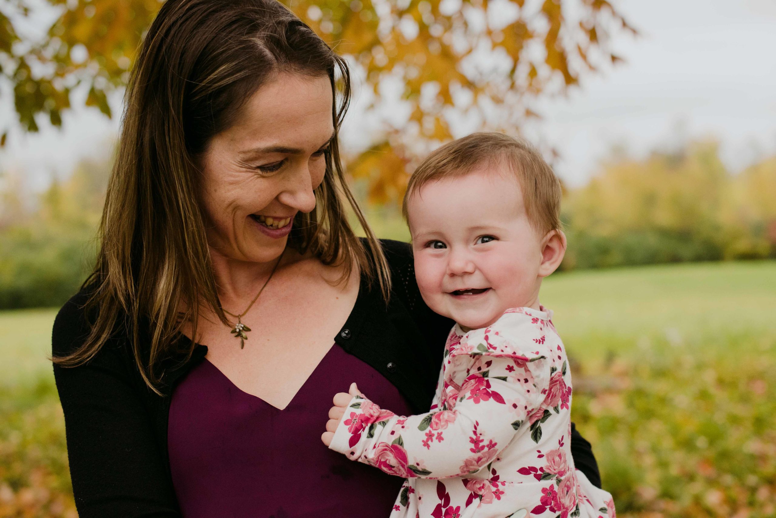 mom and baby girl smiling and laughing by fall coloured tree