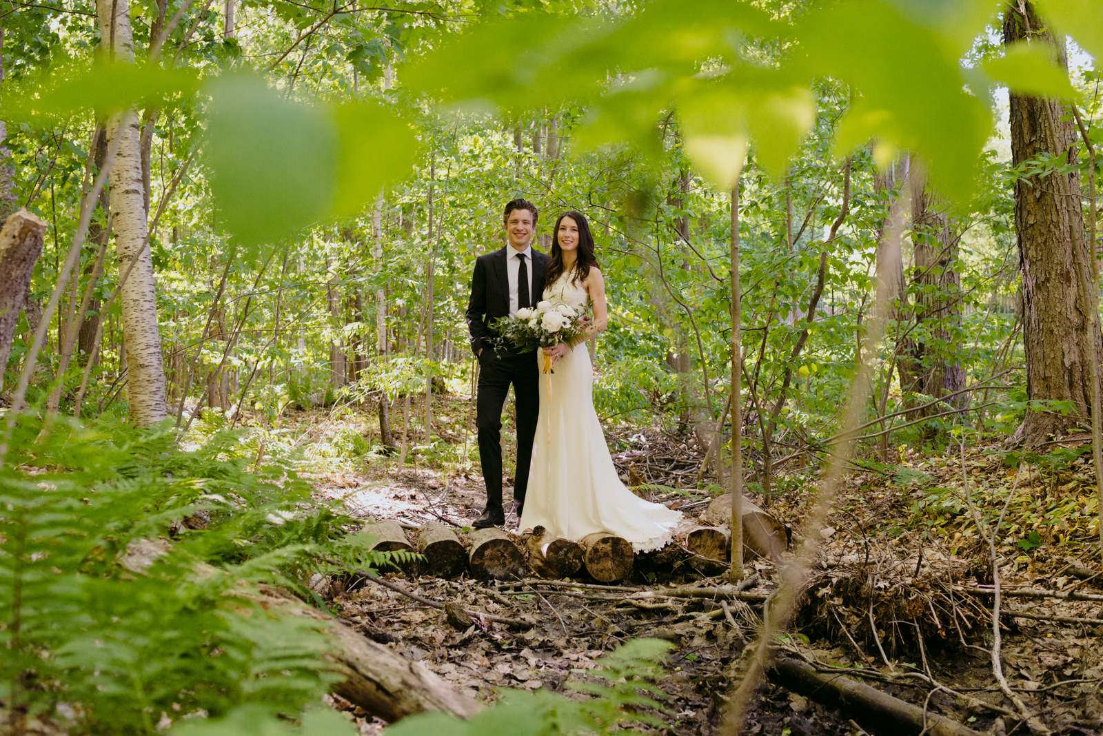 bride and groom standing on wooden logs in a forest smiling at the camera