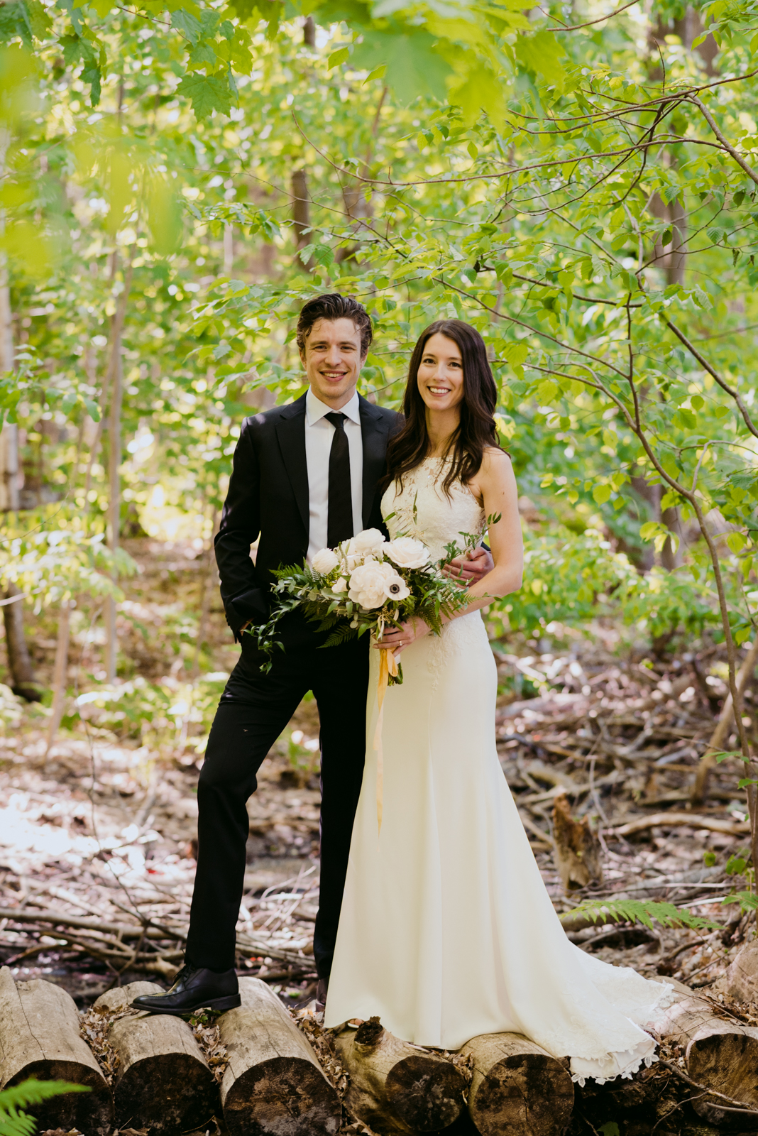bride and groom standing on wooden logs in a forest smiling at the camera