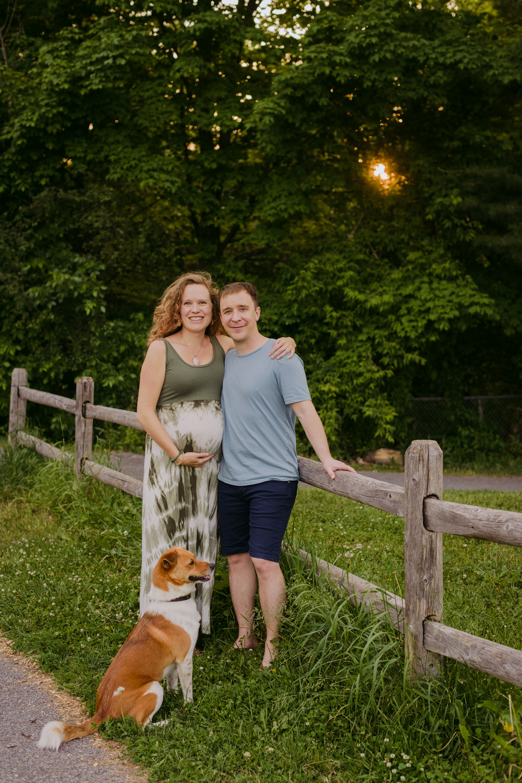 expecting parents along a wooden fence at sunset with their dog