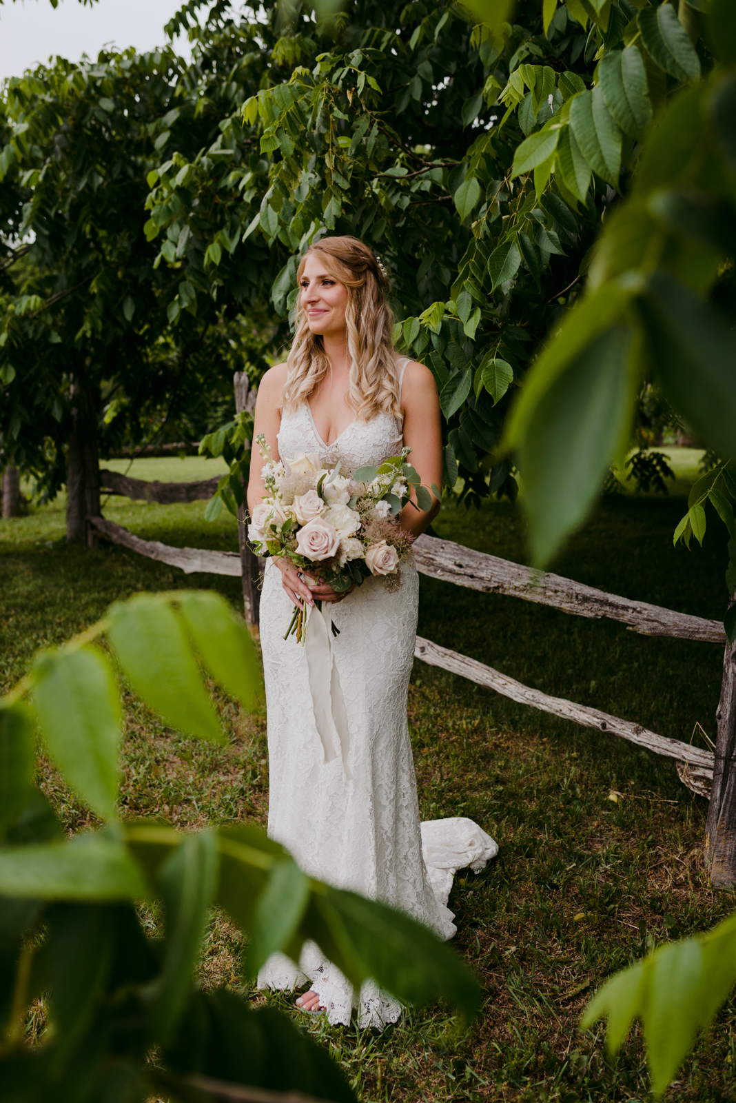 bride standing in the trees holding bouquet by wooden fence