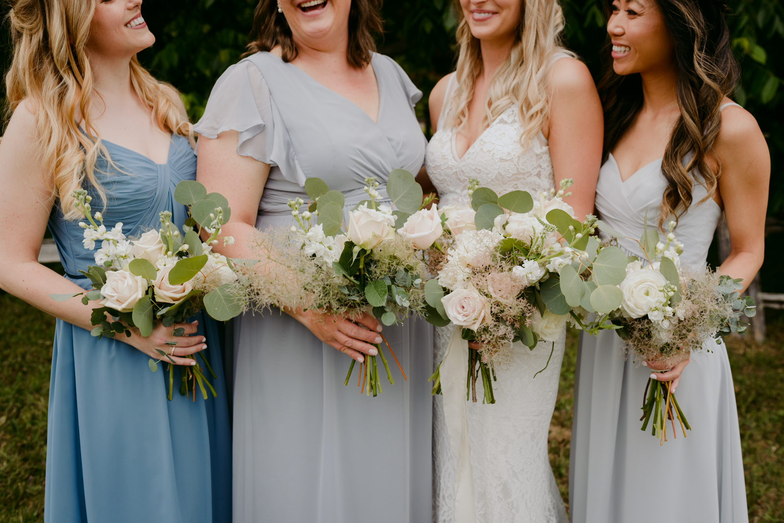 bride and bridesmaids in blue and grey holding bouquets underneath green tree