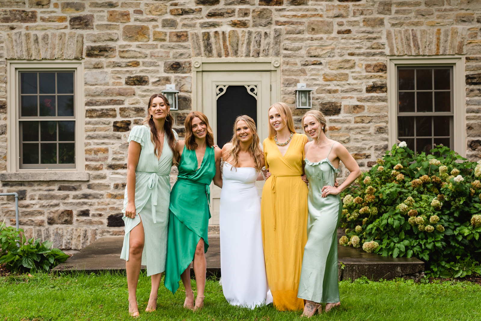 bride and bridesmaids smiling at the camera in front of old stone farmhouse