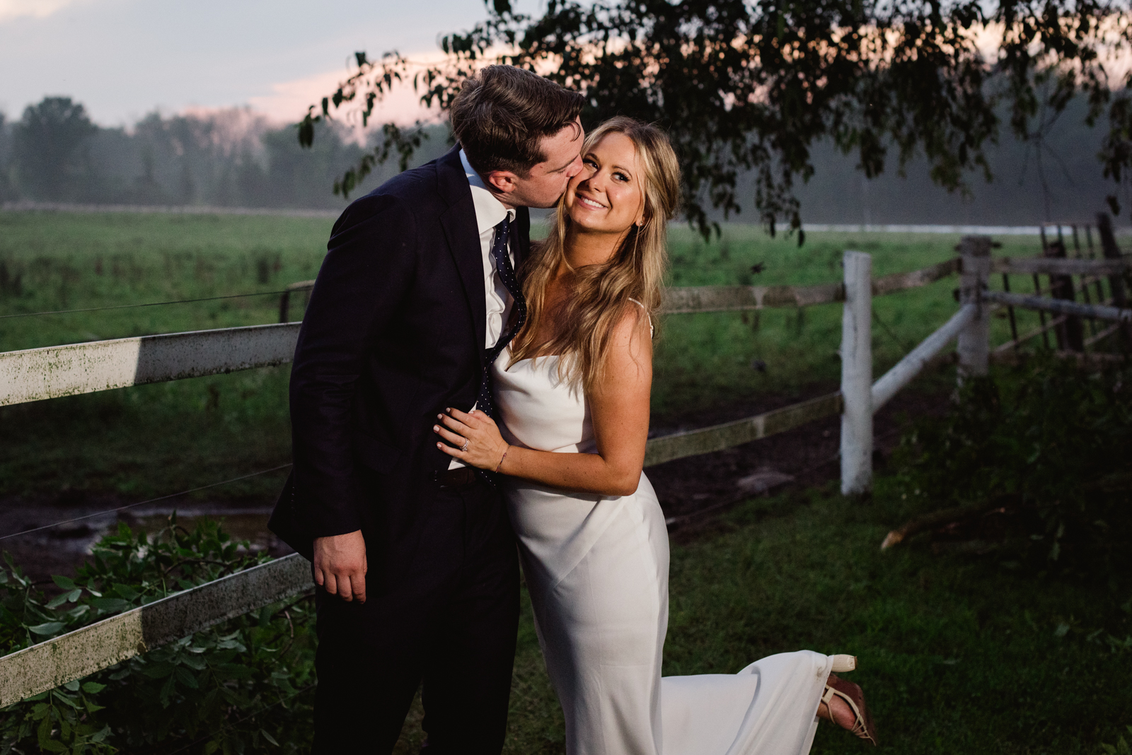 bride and groom kissing in cow's pasture at sunset
