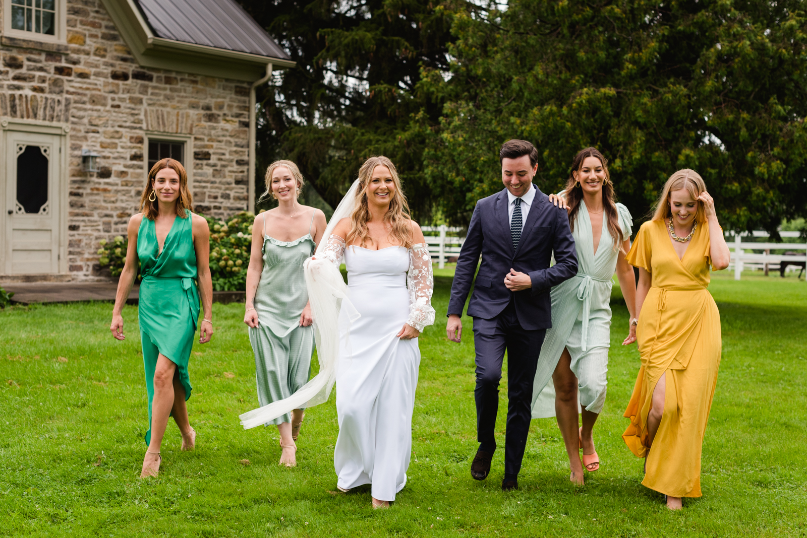 bride and bridal party walking through the grass in front of old stone farmhouse