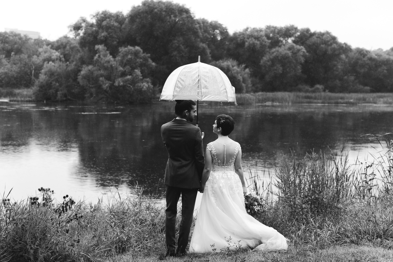 bride and groom standing by the water holding an umbrella on a rainy wedding day