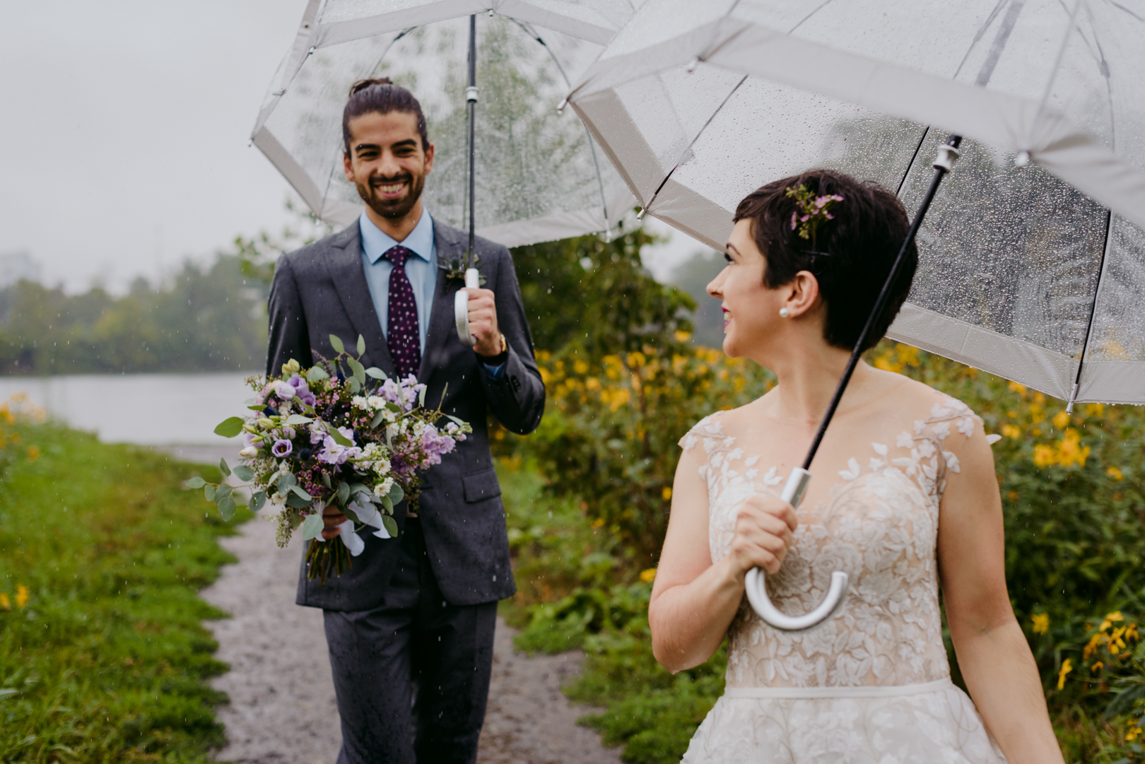 groom holding bouquet of flowers walking in the rain with umbrellas