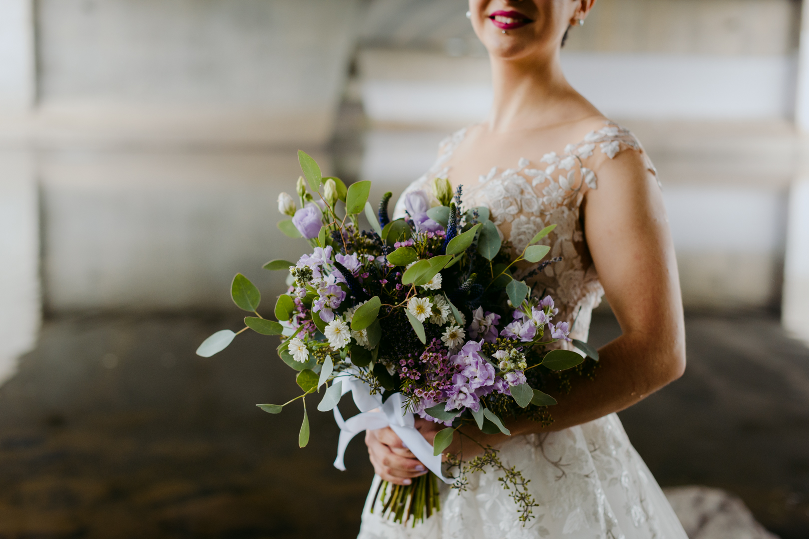 bride holding purple and green bouquet underneath a bridge by the water