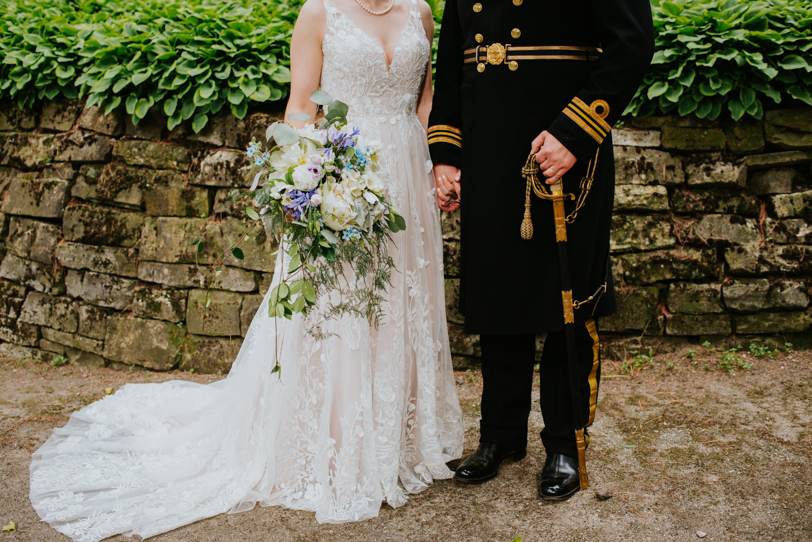 bride and groom in canadian navy uniform with sword holding hands in front of stone wall
