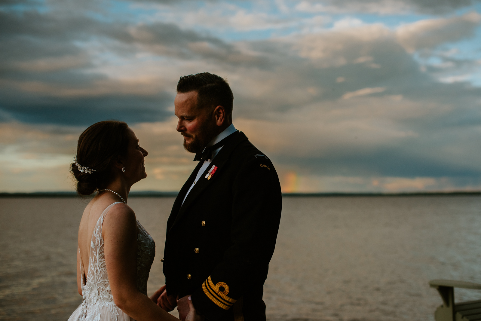 bride and groom wearing navy uniform by the water at sunset with a rainbow